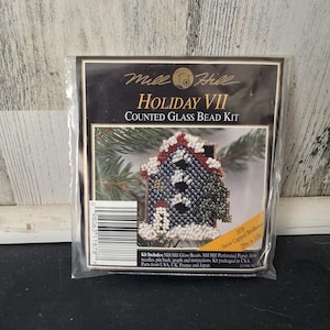 May include: A Mill Hill Holiday VII Counted Glass Bead Kit in clear plastic packaging. The package features a graphic of a dark blue house with white snow-capped roofs and a green tree. Text includes Mill Hill, Holiday VII, and Snow Capped Birdhouse.
