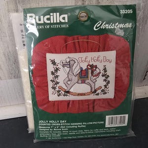 May include: A Bucilla Jolly Holly Day counted cross-stitch hanging pillow kit in plastic packaging. The kit features a rocking horse design with the text Jolly Holly Day, surrounded by a red fabric ruffle. The package states it measures 7 by 5 inches.
