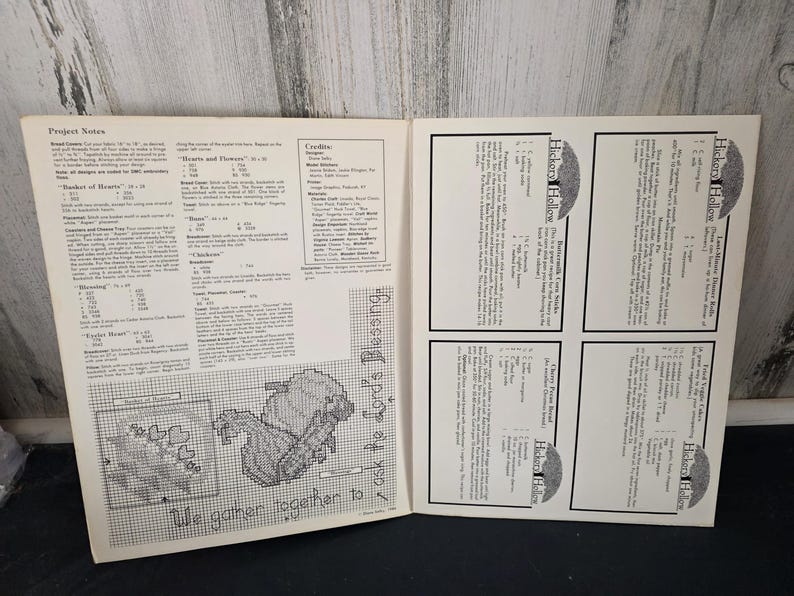 May include: Open book with pages featuring cross-stitch patterns and project notes. The text "We gather together to ask the Lord's Blessing" is visible. The book also includes the text "Hickory Hollow".