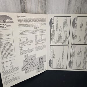 May include: An open book featuring cross-stitch patterns from Hickory Hollow. The left page includes the company's contact information and credits. The right page displays several recipe cards. The pages are white with black text and illustrations.