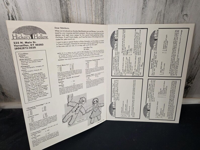 May include: An open book featuring cross-stitch patterns from Hickory Hollow. The left page includes the company's contact information and credits. The right page displays several recipe cards. The pages are white with black text and illustrations.