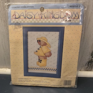 May include: A counted cross-stitch kit featuring a framed design of a bear wearing a yellow raincoat and hat. The kit includes a plastic frame and white 14 count Aida cloth. The packaging is yellow and blue, with the text "DAISY KINGDOM" and "SLICKER BEAR 5"x7".