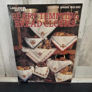 Op de afbeelding: Boekomslag met de titel "Beary Tempting Bread Cloths" met geborduurde brooddoeken met teddybeerontwerpen. De doeken worden tentoongesteld in manden en op planken, met de tekst "Leisure Arts" bovenaan.