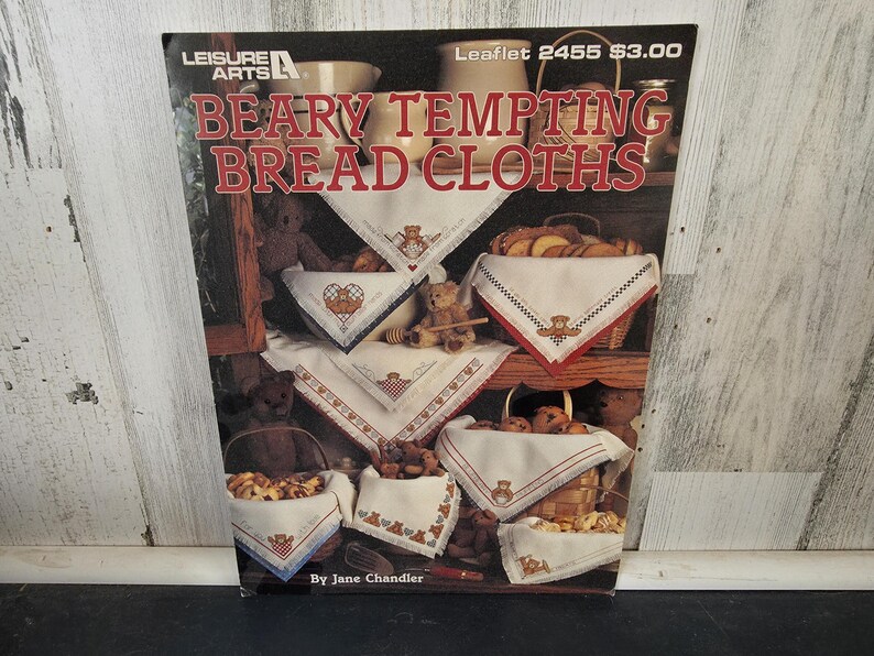 Op de afbeelding: Boekomslag met de titel "Beary Tempting Bread Cloths" met geborduurde brooddoeken met teddybeerontwerpen. De doeken worden tentoongesteld in manden en op planken, met de tekst "Leisure Arts" bovenaan.
