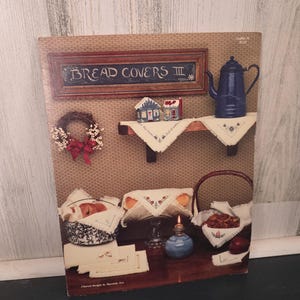 May include: A vintage print featuring a rustic kitchen scene. The print includes a framed sign that reads "BREAD COVERS III", a blue teapot, a wreath, and various baskets and linens. The color palette is warm, with browns, blues, and reds.