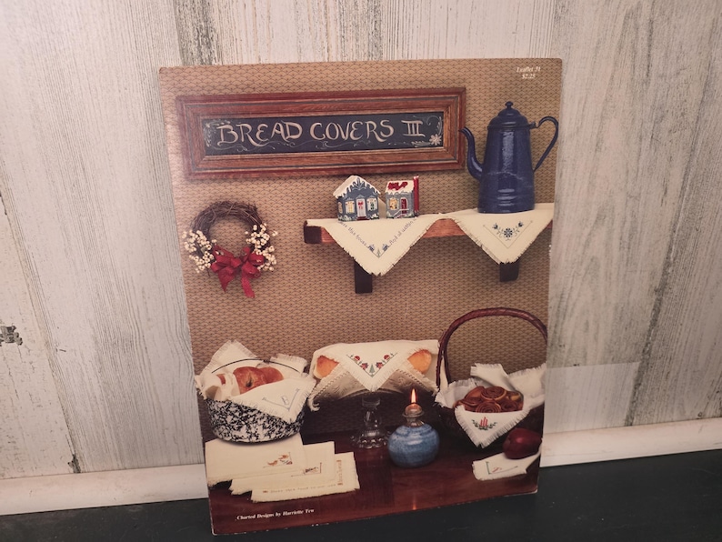 May include: A vintage print featuring a rustic kitchen scene. The print includes a framed sign that reads "BREAD COVERS III", a blue teapot, a wreath, and various baskets and linens. The color palette is warm, with browns, blues, and reds.