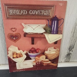Op de afbeelding: Een vintage illustratie met een rood-witte geruite achtergrond, met keukenartikelen. De afbeelding bevat een houten bord met de tekst "BREAD COVERS", een paarse theepot, een hartvormige decoratie en manden met brood en appels.