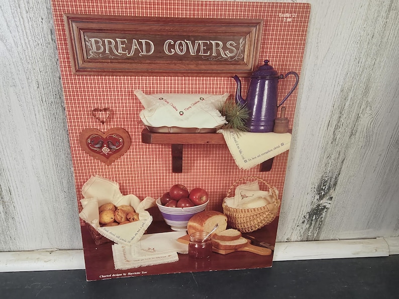 Op de afbeelding: Een vintage illustratie met een rood-witte geruite achtergrond, met keukenartikelen. De afbeelding bevat een houten bord met de tekst "BREAD COVERS", een paarse theepot, een hartvormige decoratie en manden met brood en appels.