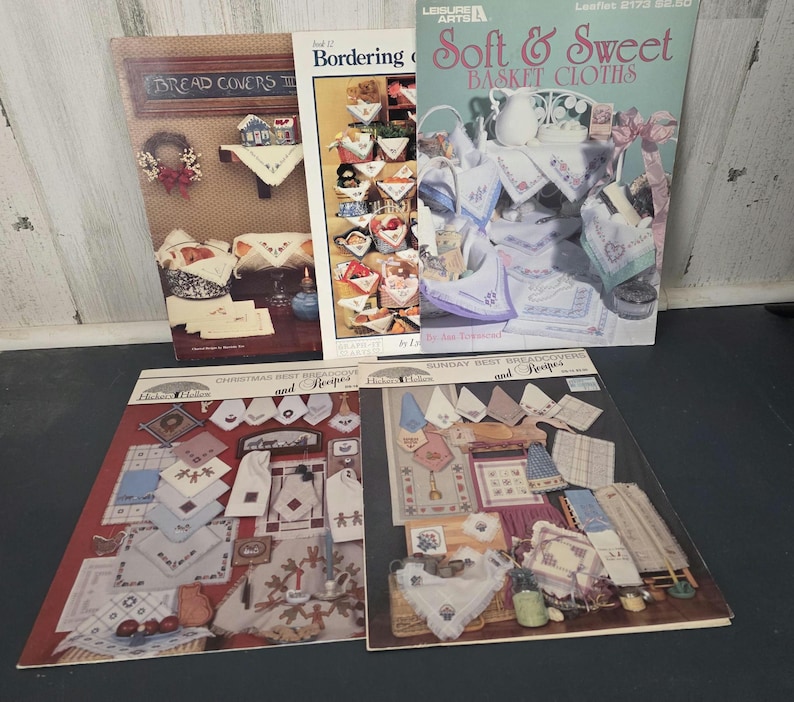 May include: A collection of vintage craft pattern books. Titles include "Bread Covers," "Bordering," "Soft & Sweet Basket Cloths," "Christmas Best Breadcovers and Recipes," and "Sunday Best Breadcovers and Recipes." The covers feature images of embroidered and crocheted items.