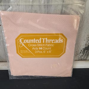 May include: A package of pink cross-stitch fabric. The label reads "Counted Threads" and includes details like "Aida 14 Count" and "3 Pcs. 6" x 6"". The fabric is all cotton and is packaged in a clear plastic bag.