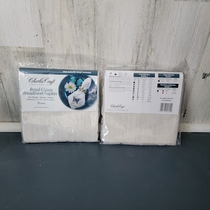 May include: Two packaged cross-stitch kits for a bread cover/napkin. The packaging features a design of a butterfly napkin ring on a plate. The text on the package reads "Royal Classic Breadcover/Napkin". The kits include instructions and a cross-stitch key.