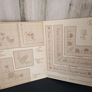 May include: Open book with cross-stitch patterns, including birds, flowers, and a house design. Text reads "Bless this house" and "And all within it." The patterns are in shades of brown and beige.