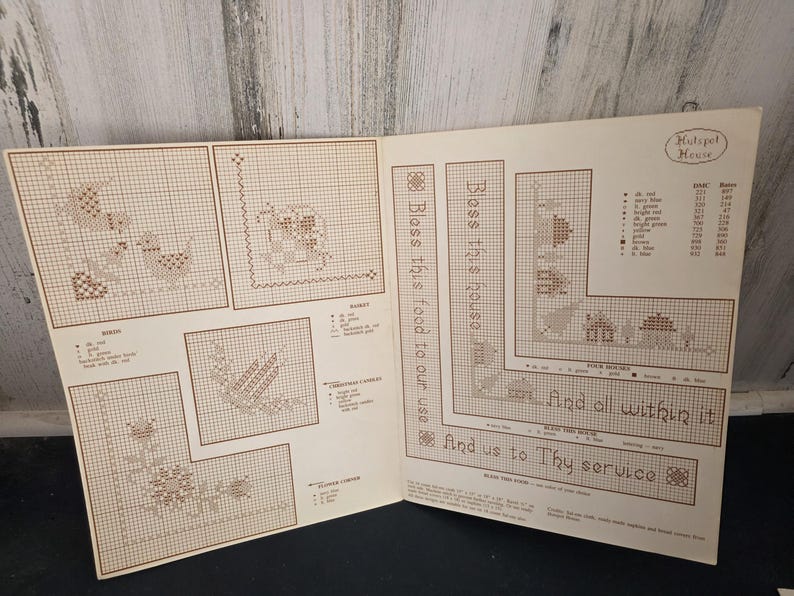 May include: Open book with cross-stitch patterns, including birds, flowers, and a house design. Text reads "Bless this house" and "And all within it." The patterns are in shades of brown and beige.