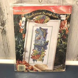 May include: A cross-stitch kit titled "Rainforest Butterflies" featuring a framed design of colorful butterflies and foliage. The kit includes Aida fabric, DMC floss, a full-color chart, and instructions. The packaging is clear plastic.