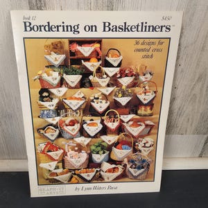 May include: A book titled "Bordering on Basketliners" with the text "36 designs for counted cross stitch." The cover displays various cross-stitch designs of baskets filled with items like fruit, flowers, and teddy bears. The author's name is Lynn Waters Busa.