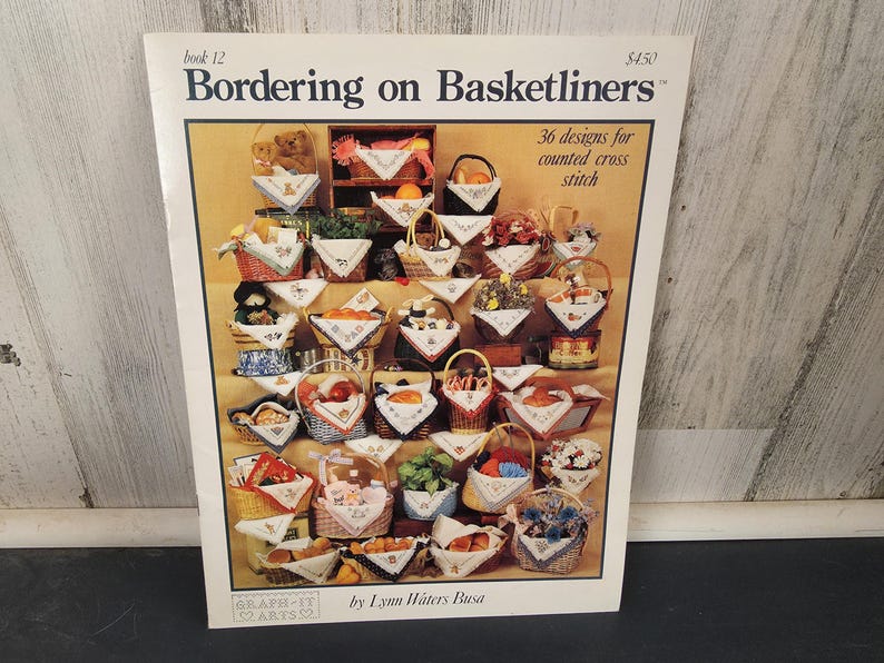 May include: A book titled "Bordering on Basketliners" with the text "36 designs for counted cross stitch." The cover displays various cross-stitch designs of baskets filled with items like fruit, flowers, and teddy bears. The author's name is Lynn Waters Busa.
