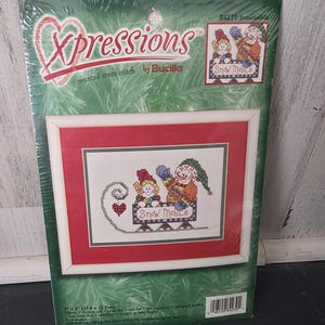 May include: A Bucilla Xpressions counted cross stitch kit featuring a snowman and a small figure riding a snowmobile. The package displays a finished 7 by 5 inch project with a red mat and white frame. The design includes a heart and text.