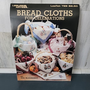 May include: A leaflet titled "Bread Cloths for Celebrations" by Leisure Arts. The cover features various baskets filled with food and decorated with embroidered cloths. The baskets contain items like eggs, nuts, and cookies. The leaflet is by Sue McElhaney.