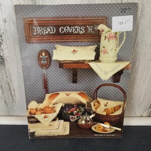 Op de afbeelding: Een display met broodhoezen, een bloemenkan en diverse bakwaren. Het bord luidt "Bread Covers II". De sc&egrave;ne omvat een plank met een broodhoes en kan, een broodmand en een kleine tafel met een theepot en fruit. Het kleurenpalet is zacht en uitnodigend.