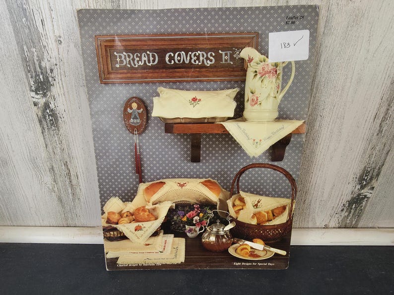 Op de afbeelding: Een display met broodhoezen, een bloemenkan en diverse bakwaren. Het bord luidt "Bread Covers II". De sc&egrave;ne omvat een plank met een broodhoes en kan, een broodmand en een kleine tafel met een theepot en fruit. Het kleurenpalet is zacht en uitnodigend.