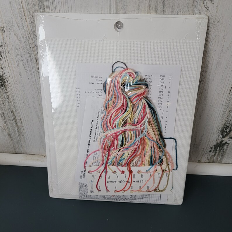 May include: A cross stitch kit featuring a white Aida cloth, colourful embroidery floss, and a pattern for a blanket with a floral design. The pattern includes a colour key and instructions for counted cross stitch.
