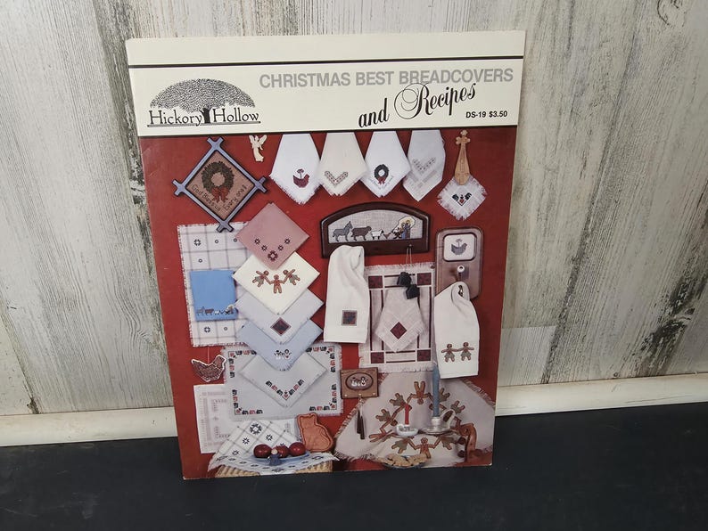 May include: A book cover titled "Christmas Best Breadcovers and Recipes" by Hickory Hollow. The cover is red with various embroidered Christmas-themed designs, including wreaths, snowflakes, and gingerbread people. The book is a collection of bread cover patterns and recipes.