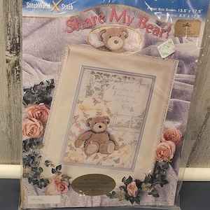 May include: A cross-stitch kit titled "Share My Bear?" featuring a framed image of a teddy bear. The kit includes fabric, cotton floss, a needle, and instructions. The frame size is 13.5 x 17.5 inches.