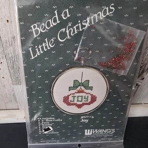 May include: A Christmas ornament kit with the text "Bead a Little Christmas". The kit includes a gold frame, floss, and a needle. The ornament design features a green bow and the word "JOY" in red. A small bag of red and green beads is included.