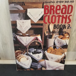 Op de afbeelding: Een vintage kookboekcover getiteld "Bread Cloths Book 2" door Carol E. Mer. De cover bevat afbeeldingen van brood, manden en geborduurde doeken. De achtergrond is een houten plank met diverse keukenartikelen.