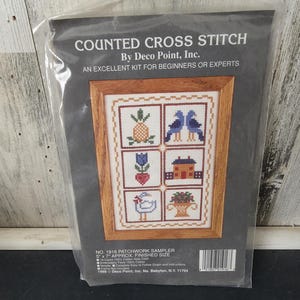 May include: A counted cross stitch kit by Deco Point, Inc. titled Patchwork Sampler. The packaging features a sampler design with six squares showing a pineapple, two blue birds, a tulip, a house, a goose, and a basket, with a 5" x 7" finished size.