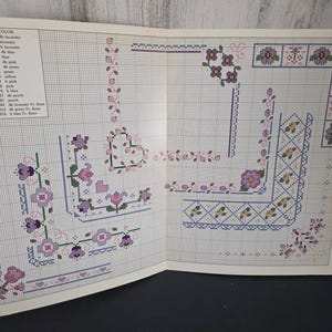 May include: Open book with cross-stitch patterns featuring floral and geometric designs in shades of purple, pink, and green. The patterns include borders, corners, and floral motifs. A color chart with thread numbers and colors is visible.
