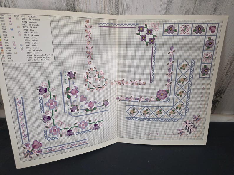 May include: Open book with cross-stitch patterns featuring floral and geometric designs in shades of purple, pink, and green. The patterns include borders, corners, and floral motifs. A color chart with thread numbers and colors is visible.