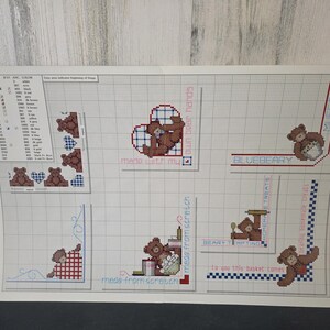 Op de afbeelding: Een kruissteekpatroonvel met teddybeerontwerpen. De vel bevat verschillende teddybeermotieven, harten en tekstelementen zoals "made with my own bear hands" en "blueberry treats". Het kleurenpalet omvat bruin, rood en blauw.