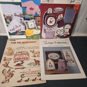 May include: A collection of five vintage cross-stitch pattern booklets featuring various designs including wildlife, Christmas themes, and decorative home motifs. The covers display finished embroidery projects with text such as For The Sportsman, The Artists Collection, and Hickory Hollow.