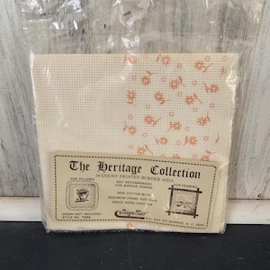 May include: A packaged cross-stitch kit from The Heritage Collection. The kit includes a cream-colored Aida cloth with a floral pattern in a light orange hue. The label indicates it is for pillows and framing.