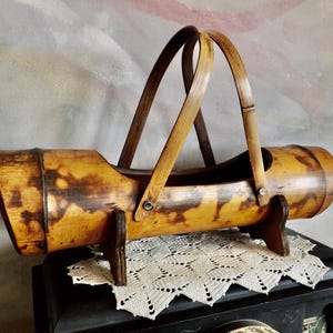 May include: A vintage bamboo wine bottle holder with a natural finish. The holder features a cylindrical body with a handle and is supported by two wooden feet. The bamboo has dark brown markings.