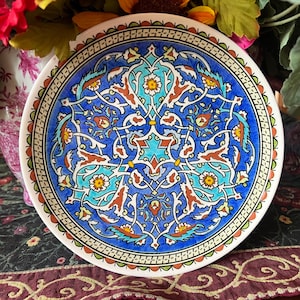 May include: A round ceramic decorative plate featuring a vibrant blue background with intricate, symmetrical floral and vine patterns in shades of turquoise, white, orange, and yellow, finished with a detailed geometric border along the rim.