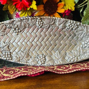 May include: A rectangular silver-toned metal tray featuring a central woven basket-weave pattern. The border is decorated with a raised relief design of grape clusters and grape leaves that extends around the entire perimeter of the tray.