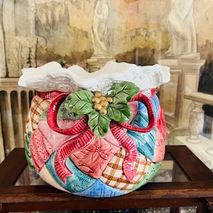 May include: A ceramic bowl featuring a quilted patchwork pattern in shades of red, blue, green, and tan. The bowl has a white ruffled rim and a central decorative element consisting of green holly leaves and gold berries tied with a red ribbon.