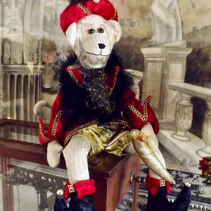 May include: A plush monkey doll wearing a red and gold costume with a black feathered hat. The monkey is sitting on a wooden surface with a glass case behind it.