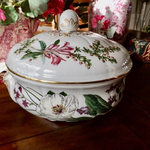 Puede incluir: Una fuente de cerámica blanca con tapa, con diseños florales y botánicos en rosa, verde y morado. La fuente tiene un borde dorado y un pomo decorativo. Una pieza clásica para servir alimentos.