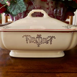 May include: Cream-colored, lidded serving dish with a decorative black floral design. The dish has a rectangular shape with a slightly curved lid and a handle. A red trim accents the rim.