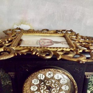 May include: An antique gold-colored ornate picture frame with a small image inside, resting on a black clock. The clock face is gold with black Roman numerals. The frame has a detailed, scroll-like design.