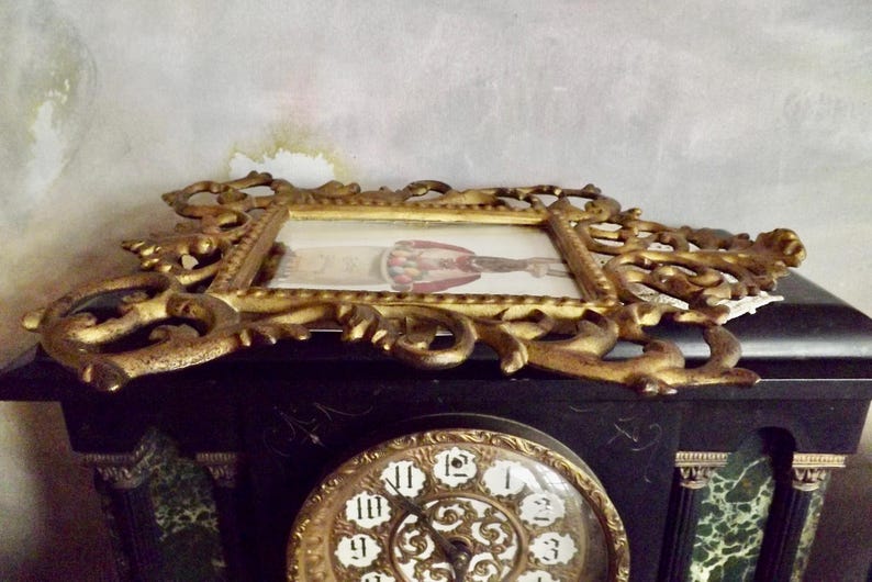 May include: An antique gold-colored ornate picture frame with a small image inside, resting on a black clock. The clock face is gold with black Roman numerals. The frame has a detailed, scroll-like design.