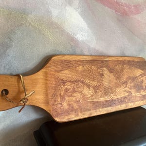 May include: A rectangular wooden serving board with a handle featuring a hole and a leather cord. The surface displays a detailed laser-engraved image of a bald eagle perched on a branch with leaves, showcasing natural wood grain and warm brown tones.