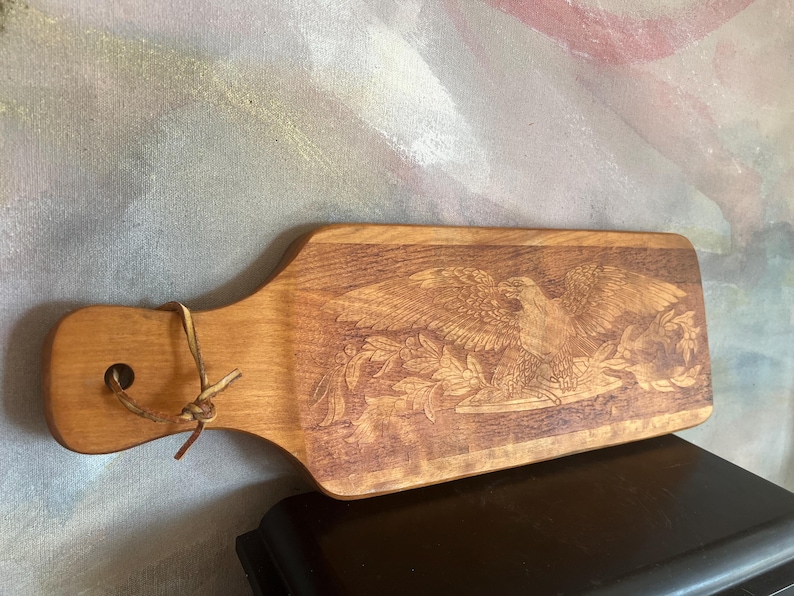 May include: A rectangular wooden serving board with a handle featuring a hole and a leather cord. The surface displays a detailed laser-engraved image of a bald eagle perched on a branch with leaves, showcasing natural wood grain and warm brown tones.
