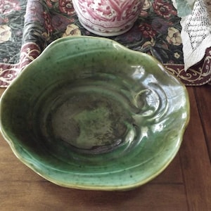 May include: A handcrafted, green ceramic bowl with an organic, wavy rim. The bowl has a glossy finish and a dark interior, suggesting a unique, artisanal piece. The bowl is likely used for serving or display.