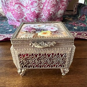 May include: A rectangular metal jewelry box with a gold-tone finish, featuring an ornate filigree pattern over a deep red lining. The lid displays a painted floral design of white, pink, and yellow roses, finished with a decorative metal latch.