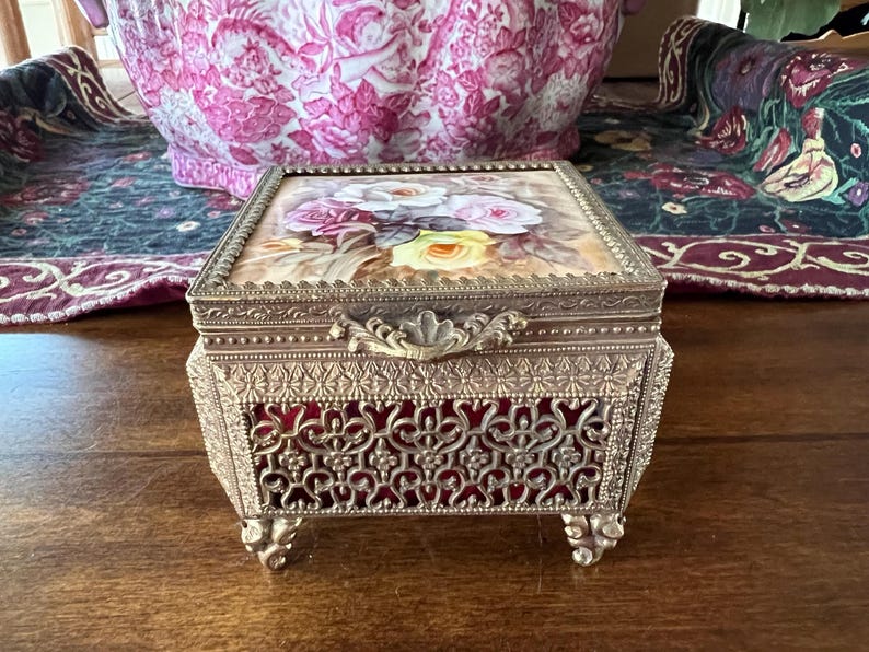 May include: A rectangular metal jewelry box with a gold-tone finish, featuring an ornate filigree pattern over a deep red lining. The lid displays a painted floral design of white, pink, and yellow roses, finished with a decorative metal latch.