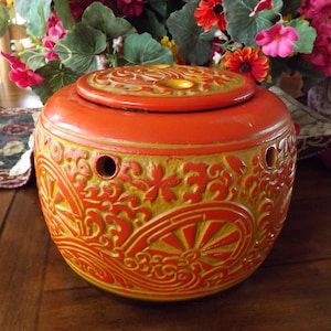 May include: A round, orange ceramic container with a lid, featuring a detailed yellow floral and geometric relief pattern. The container has several small holes and sits on a wooden surface, with flowers in the background.
